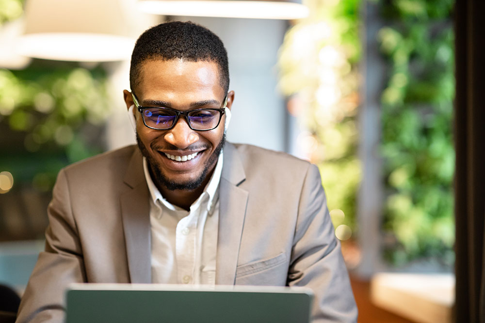 Male Executive. Happy black businessman using laptop, listening to music in wireless headphones, sitting at cafe, copyspace